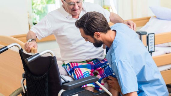 Elderly care nurse in retirement home helping senior from wheel chair to bed